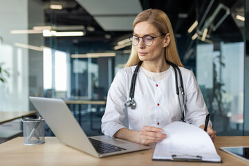 Professional female doctor in white coat with stethoscope working on laptop and examining medical documents. Scene conveys focus, expertise, and concentration in healthcare and medical field.