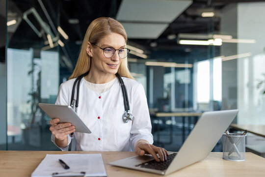 Professional female doctor working in bright office using laptop and tablet for virtual consultation. She wears a stethoscope and white coat, reflecting modern healthcare practices.