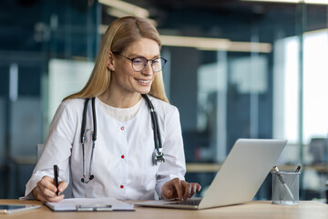 Confident female doctor wearing glasses and stethoscope using laptop in professional office setting. She is writing on paper, showcasing multitasking skills in medical industry.