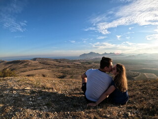 Fototapeta premium Couple sitting on a hilltop overlooking a vast mountainous landscape, sharing a kiss while enjoying the sunset under a partly cloudy sky during a peaceful outdoor moment surrounded by nature