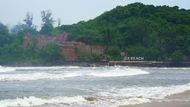 Wide shot showing shot of Baga Beach with tree covered hills in the background and brown muddy water waves rolling into the beach showing this popular tourist spot