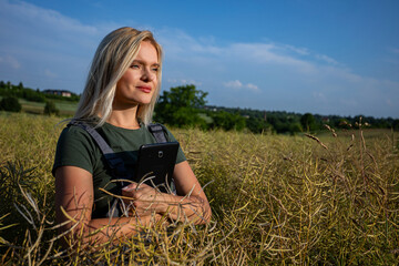 A woman farmer standing in the middle of rapeseed field, holding a tablet close to her body and satysfied with her rapeseed crop