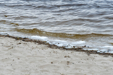 River bank with waves and white foam. White soft foam swaying. Blue water edge on sandy beach. Swirl natural pattern in shallow water. Tranquil scene of beach.