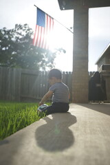 Young boy sitting on the grass with a soccer ball in the backyard, the American flag draped overhead with sunlight shining through