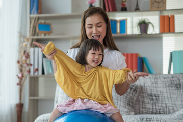 Happy Asian child having fun with fitness ball, doing exercises with mother at home