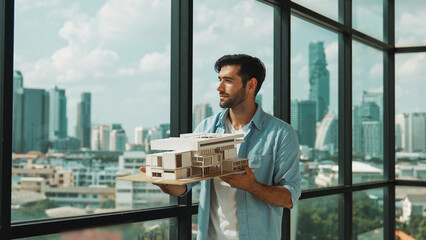 Skilled smiling caucasian engineer turns house model left and right to check mistake point. Professional manager inspect at architectural model while standing near window. Design concept. Tracery