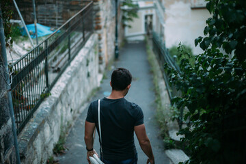 A man on a narrow street of a European city in an ordinary residential quarter of the historical center. He is wearing a grey T-shirt. The street is green, brick wall, houses. Coming down the stairs