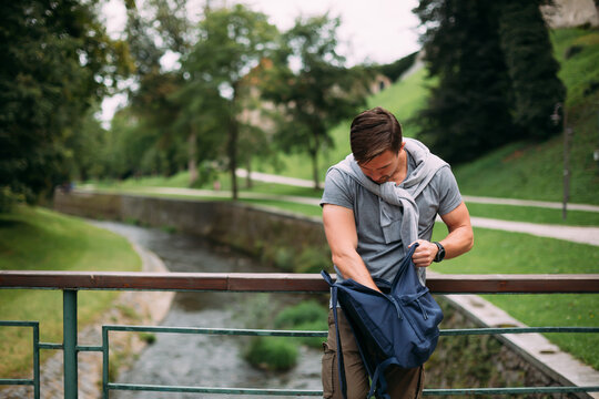 The man is looking for something in his blue backpack. The tourist is trying to find something in his backpack. A tourist stands against the background of a river and a park. The man lost an item