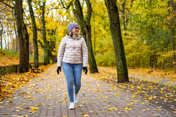 Fototapeta premium Beautiful middle-aged blonde woman wearing jeans, beige jacket and winter hat and sunglasses walking on alley full of yellow fallen leaves in park on autumn day. Front view. Autumnal walk in city park