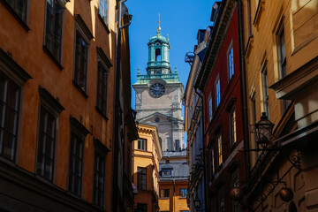 Fototapeta premium A narrow alleyway lined with beautifully aged buildings reveals a charming clock tower of St. Nicholas church or Storkyrkan in the background, bathed in sunlight on a clear day in Stockholm, Sweden.