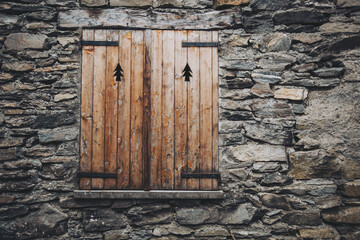 antique wooden window on a construction made of slate, typical of the villages of architecture in the province of Alps Mountains. Closed wooden window shutters with with typical alpine decoration