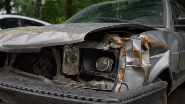 Closeup of aged abandoned car with serious damage, rust, and missing parts after impact and collision. Old vehicle left on urban street on cloudy summer day, nobody. Tracking shot in slow motion.