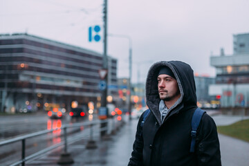 There is a man on the street. The weather is rainy. There are lights of stop signals. A man wears a black winter jacket. There are buildings on the background