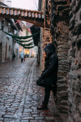 A man in a black hooded jacket on a cobblestone street in an ancient city. A narrow European street. The man is leaning against the wall, the hood is on, the face is not visible