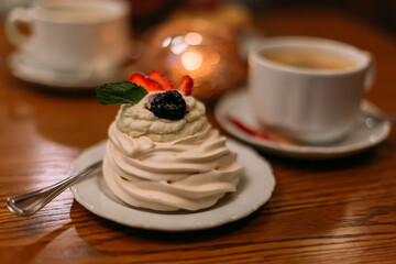 Delicious Pavlova Dessert with Whipped Cream and Berries, Served with a Cup of Coffee in a Cozy Café Setting, Elegant Pastry on Wooden Table, Gourmet Sweet Treat for Relaxing Moment