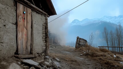 Fototapeta premium A weathered wooden door leads into a rustic stone hut, set against a backdrop of snow-capped mountains shrouded in mist. The scene evokes a sense of tranquility, isolation, and the enduring nature of 