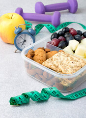 Healthy snack, diet, fitness, weight control. Lunch box with a light snack, cereal cookies, fruits, berries, nuts, dumbbells and tape measure on a light background.