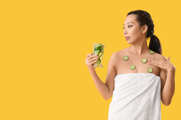 Young Asian woman with glass of cucumber water on yellow background