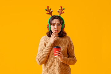 Worried young woman in reindeer horns with cup of coffee on yellow background