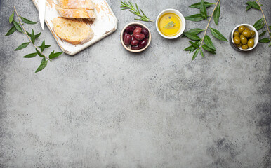 Composition with sliced fresh ciabatta on cutting board, green and brown olives, olive oil with rosemary, olive tree branches on gray concrete stone rustic background top view, copyspace border