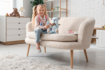 Obraz premium Cute little girl with first aid kit and teddy bear playing on sofa at home