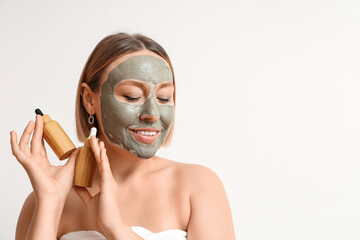 Mature woman with clay mask holding bottles of essential oil on light background