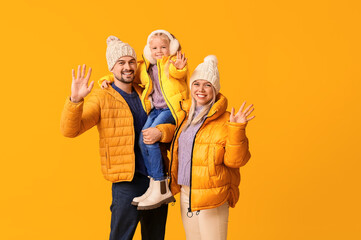 Happy couple with daughter in winter clothes waving hands on yellow background