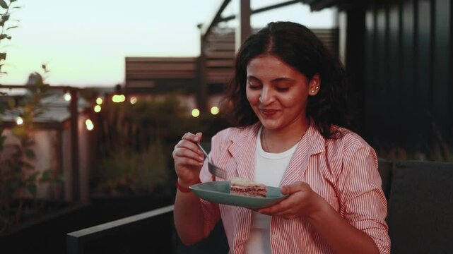 Pretty young indian woman looking around to make sure nobody watching and eating cake with great pleasure at home terrace Smiling female biting and enjoying bakery indoors Break the diet concept