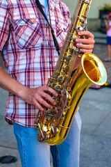 Obraz premium A talented street musician is playing a golden saxophone while standing on a city street corner. He is wearing a casual outfit and has a strap around his neck to hold the instrument.