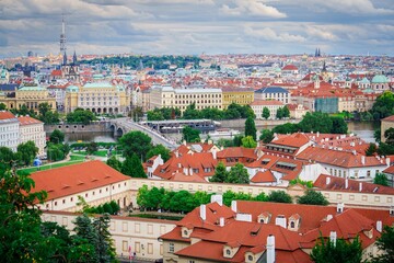 Naklejka premium A beautiful view of Prague featuring red-roofed buildings along the Vltava River. Iconic bridges and green parks are set against a clear blue sky with fluffy white clouds, creating a stunning