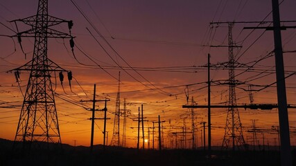 Fototapeta premium Landscape photograph featuring a sunset. The layout includes numerous power lines and transmission towers of varying heights and designs, creating a complex network of intersecting lines. 