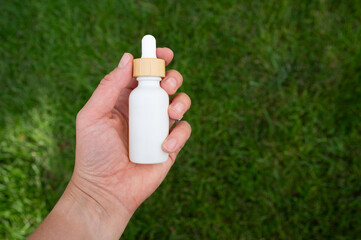 Hand holding a white oil dropper bottle against green grass background.