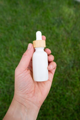 Hand holding a white oil dropper bottle against green grass background.