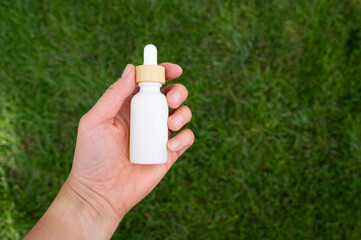 Hand holding a white oil dropper bottle against green grass background.