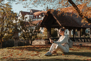 Obraz premium Young Fashionable Teenage Girl With Smartphone In Europian Park In Autumn Sitting At Smiling. Trendy Young Woman In Fall In Park Texting. Retouched, Vibrant Colors. Beautiful Blonde Teenage Girl