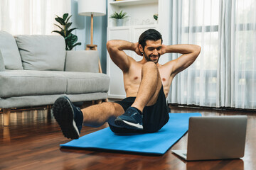 Athletic and sporty man doing crunch on fitness mat while follow online home workout exercise instruction for fit physique and healthy sport lifestyle at home. Online gaiety home exercise video.