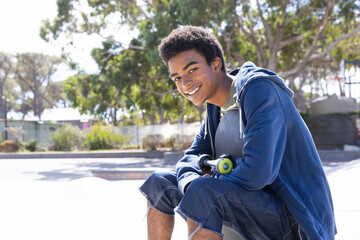 Smiling african american teenage boy sitting with skateboard in park, enjoying leisure time outdoors