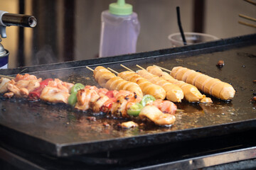 Close-up of sausages and skewered meat being grilled on a hotplate, with a smoky, sizzling effect capturing the essence of street food preparation
