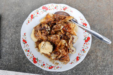 Close-up of Batagor, a traditional Indonesian street food from Bandung, served on a floral-patterned plate with peanut sauce and sweet soy sauce. A spoon rests beside the crispy fried dumplings