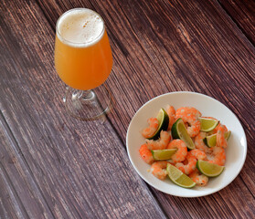A tall glass of light wheat beer and a plate of boiled shrimp with slices of lime on a wooden table.