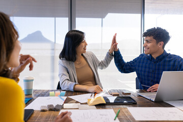 High-fiving, diverse colleagues celebrating success with laptop and design materials in office
