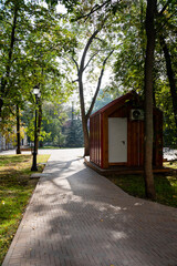 A charming and quaint small wooden shed is beautifully situated right beside a winding path that runs through a picturesque park, surrounded by lush greenery and scenic nature all around it