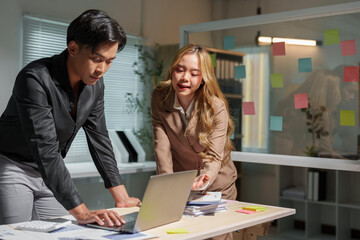 A team of business men and women wrote and pointed at sticky notes on the glass to brainstorm ideas. Propose ideas and write on the glass board. Discuss. Meeting information on a laptop in the office.