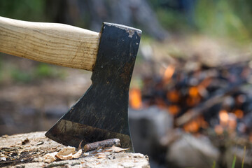 Close-up of an axe embedded in a log with a campfire burning in the background