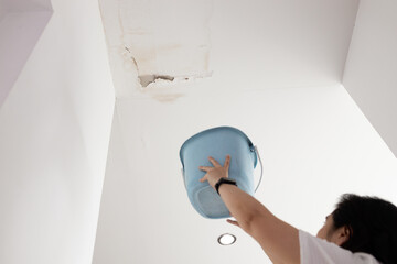 Anxious asian woman standing holding plastic bucket during heavy rain,worried about water dripping on the floor from leaking roof,dirty water stains,water leakage,damage on ceiling surface in bedroom © Satjawat