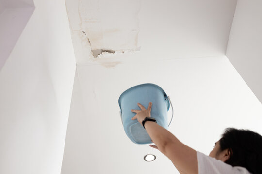 Anxious asian woman standing holding plastic bucket during heavy rain,worried about water dripping on the floor from leaking roof,dirty water stains,water leakage,damage on ceiling surface in bedroom