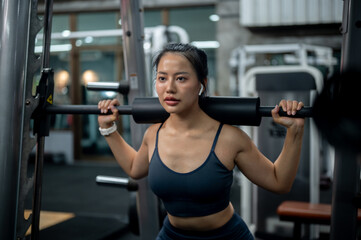 A sporty Asian woman in athletic wear is performing squats on the Smith machine at the gym.