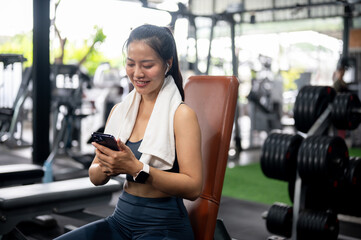 A cheerful Asian woman is using her smartphone while resting on a bench press in the gym.