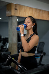 An attractive, sporty Asian woman is drinking water while walking on a treadmill in the fitness.