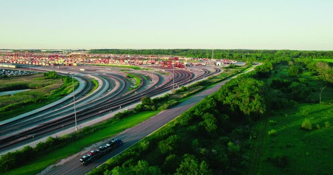 Beautiful aerial of stinger car hauler driving on a highway, parallel to railway tracks, surrounded by vibrant forest and open fields.